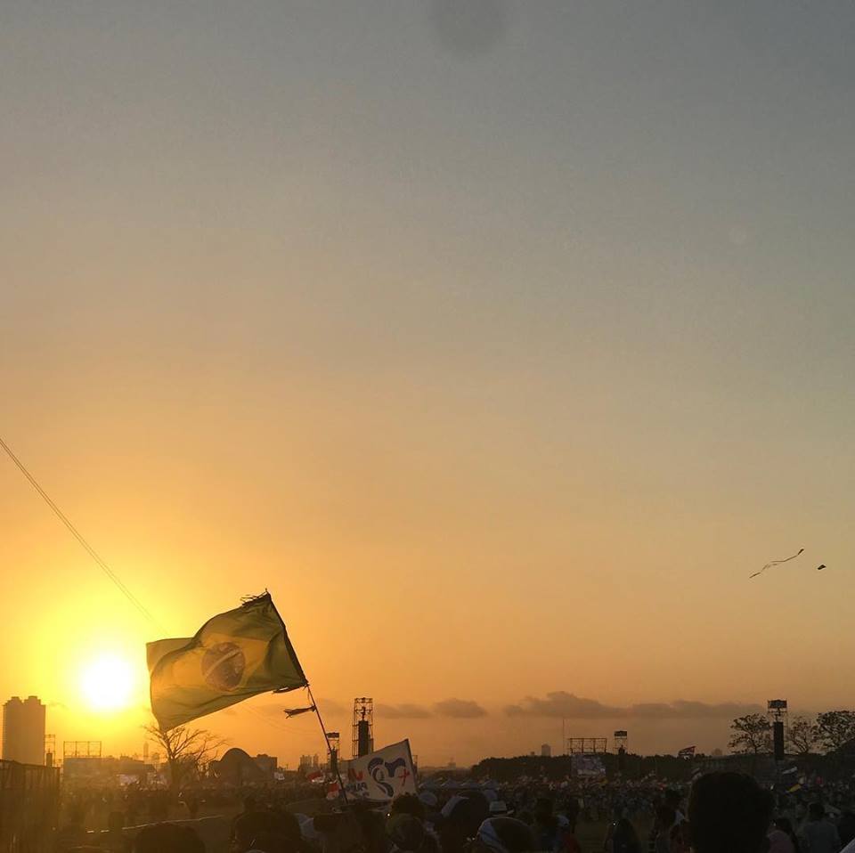A foto mostra uma bandeira do Brasil em frente ao pôr-do-sol e milhares de jovens que participaram do evento.